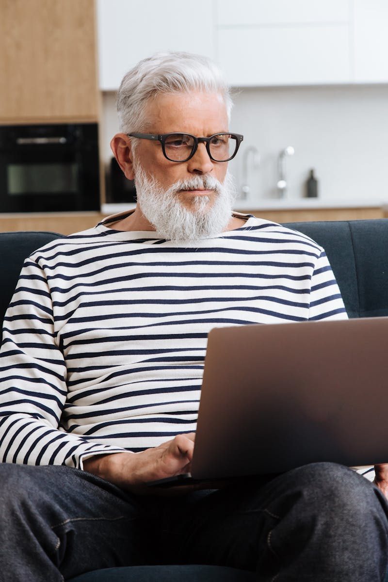 Photo by Ron Lach Elderly man with glasses focused on laptop work, sitting indoors in a cozy setting.
